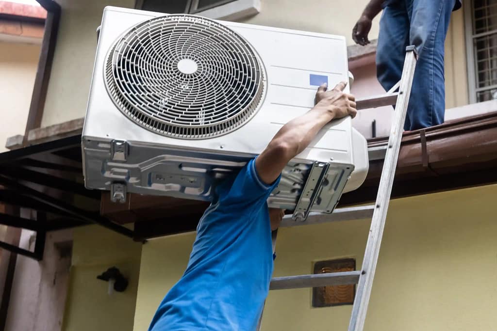 Worker carrying an air conditioning unit up a ladder for installation.