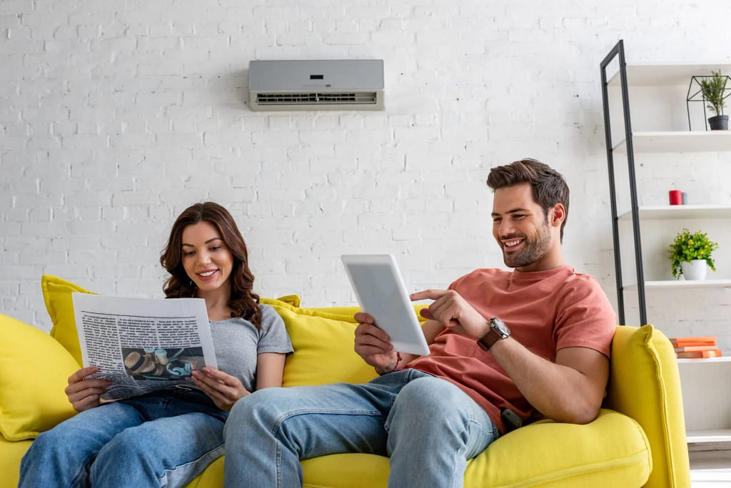 Couple relaxing on yellow couch, reading newspaper and tablet, with air conditioning unit.